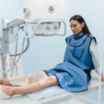 Woman in a protective lead apron sits on a treatment couch with her knee positioned under an imaging machine