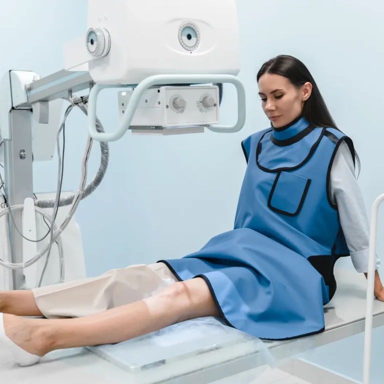 Woman in a protective lead apron sits on a treatment couch with her knee positioned under an imaging machine
